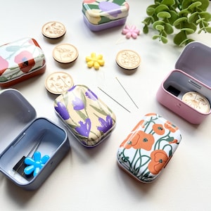 May include: Six small metal tins with floral designs. The tins are open and some contain sewing needles and a blue plastic flower. The tins are on a white surface with other small decorative items.