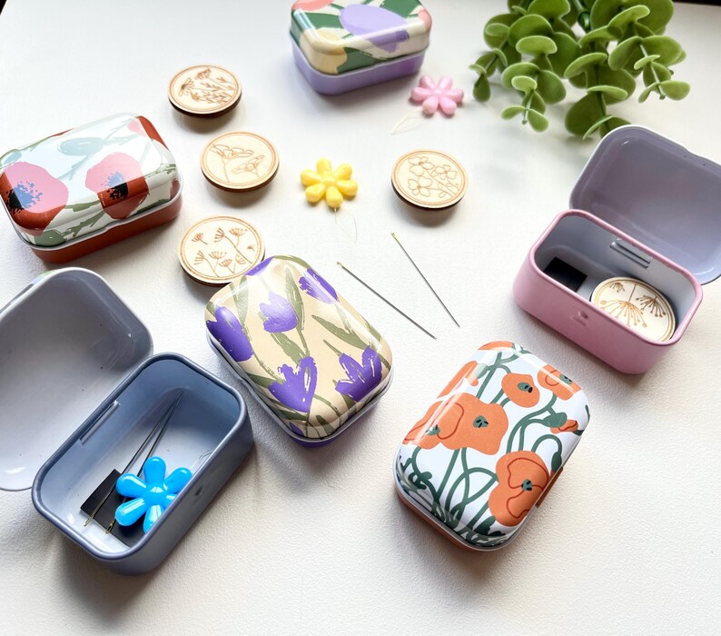 May include: Six small metal tins with floral designs. The tins are open and some contain sewing needles and a blue plastic flower. The tins are on a white surface with other small decorative items.