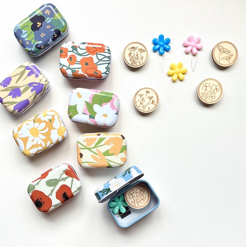 May include: Assortment of small, floral patterned metal tins with lids. Each tin contains a small wooden button with a floral design. There are also three small plastic flowers with pins attached.