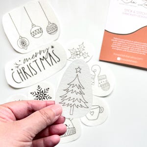 Puede incluir: Una colecci&oacute;n de dise&ntilde;os de bordado blancos, despegables y adhesivos, con ilustraciones navide&ntilde;as. Los dise&ntilde;os incluyen un &aacute;rbol de Navidad, adornos, copos de nieve, acebo, un guante, una taza y el texto "merry CHRISTMAS".