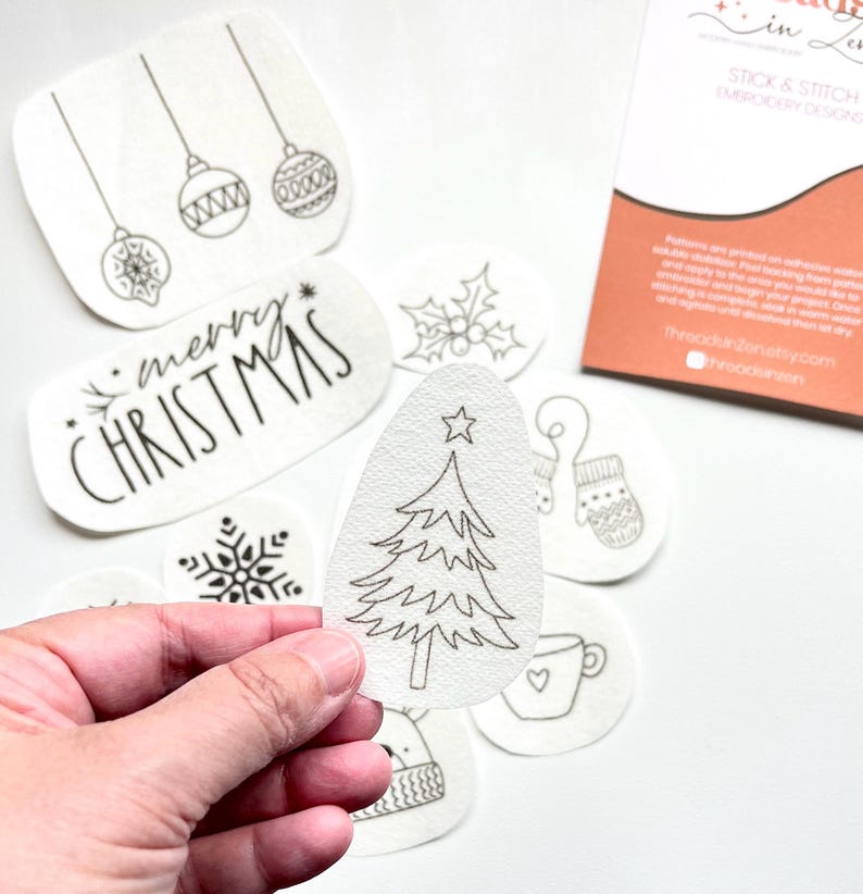 Puede incluir: Una colecci&oacute;n de dise&ntilde;os de bordado blancos, despegables y adhesivos, con ilustraciones navide&ntilde;as. Los dise&ntilde;os incluyen un &aacute;rbol de Navidad, adornos, copos de nieve, acebo, un guante, una taza y el texto "merry CHRISTMAS".