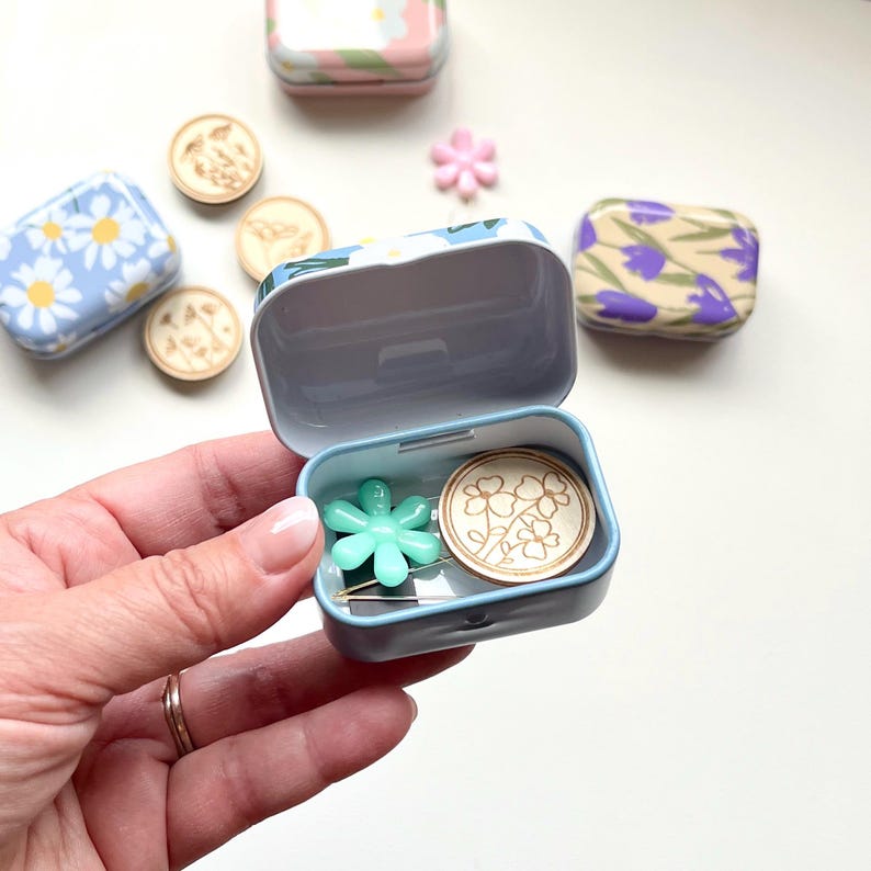 May include: A small blue and white floral tin box with a hinged lid is open and contains a wooden button with a floral design, a teal blue flower-shaped pin, and a silver pin.