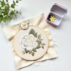 May include: A white and green floral embroidery design in a wooden embroidery hoop on a beige fabric background.