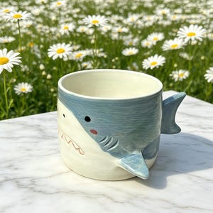 Puede incluir: Una taza de cer&aacute;mica con un dise&ntilde;o de tibur&oacute;n. La taza es principalmente blanca con un dise&ntilde;o de tibur&oacute;n azul claro en el exterior. El tibur&oacute;n tiene ojos negros, una mejilla roja y una sonrisa. El asa tiene forma de aleta de tibur&oacute;n.