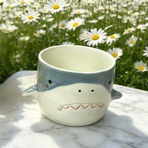Puede incluir: Taza de cer&aacute;mica con forma de tibur&oacute;n, en color azul claro y blanco. La taza presenta un dise&ntilde;o de cara de tibur&oacute;n con ojos negros, una sonrisa con dientes y peque&ntilde;as aletas. Est&aacute; sobre una superficie de m&aacute;rmol, con un fondo de margaritas.