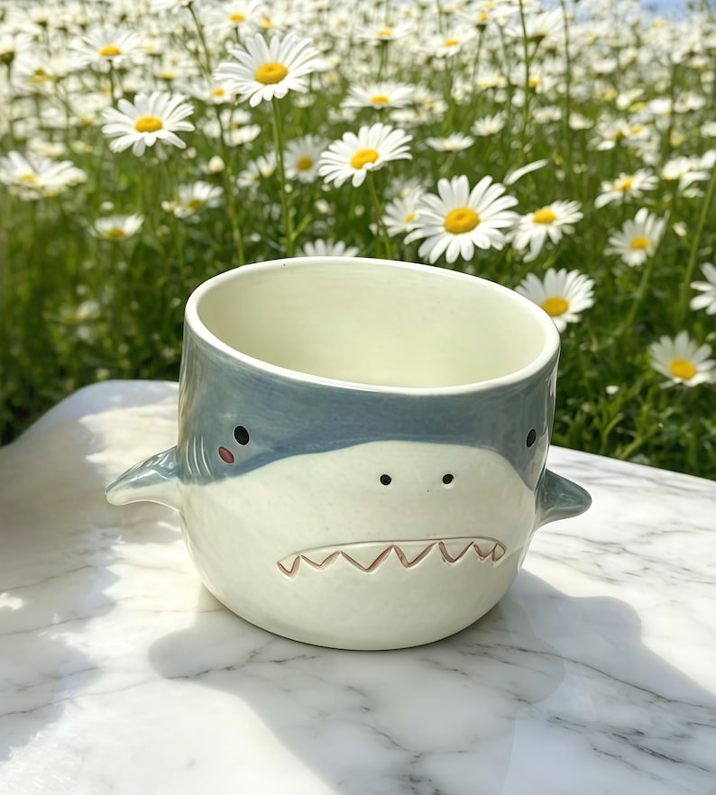 Puede incluir: Taza de cer&aacute;mica con forma de tibur&oacute;n, en color azul claro y blanco. La taza presenta un dise&ntilde;o de cara de tibur&oacute;n con ojos negros, una sonrisa con dientes y peque&ntilde;as aletas. Est&aacute; sobre una superficie de m&aacute;rmol, con un fondo de margaritas.