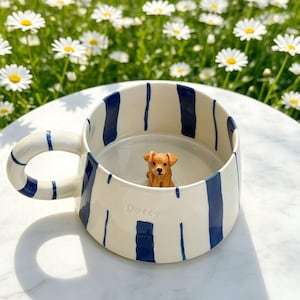 Puede incluir: Una taza de cer&aacute;mica con base blanca y rayas verticales azul marino. La taza presenta una peque&ntilde;a figura tridimensional de un perro marr&oacute;n unida al fondo interior y la palabra Dotty grabada en el lado exterior.