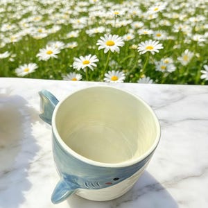 Puede incluir: Una taza de cer&aacute;mica con dise&ntilde;o de tibur&oacute;n. La taza es blanca con un asa de aleta de tibur&oacute;n azul claro y un dise&ntilde;o de cara de tibur&oacute;n azul y blanco. La taza est&aacute; sobre una superficie de m&aacute;rmol blanco.