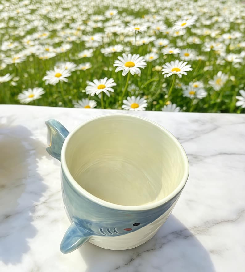 Puede incluir: Una taza de cer&aacute;mica con dise&ntilde;o de tibur&oacute;n. La taza es blanca con un asa de aleta de tibur&oacute;n azul claro y un dise&ntilde;o de cara de tibur&oacute;n azul y blanco. La taza est&aacute; sobre una superficie de m&aacute;rmol blanco.