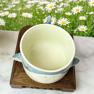 Puede incluir: Un cuenco de cer&aacute;mica con tem&aacute;tica de tibur&oacute;n con un dise&ntilde;o azul claro y blanco, con asa y aletas. El cuenco se encuentra sobre un soporte de madera oscura, con un fondo de margaritas.