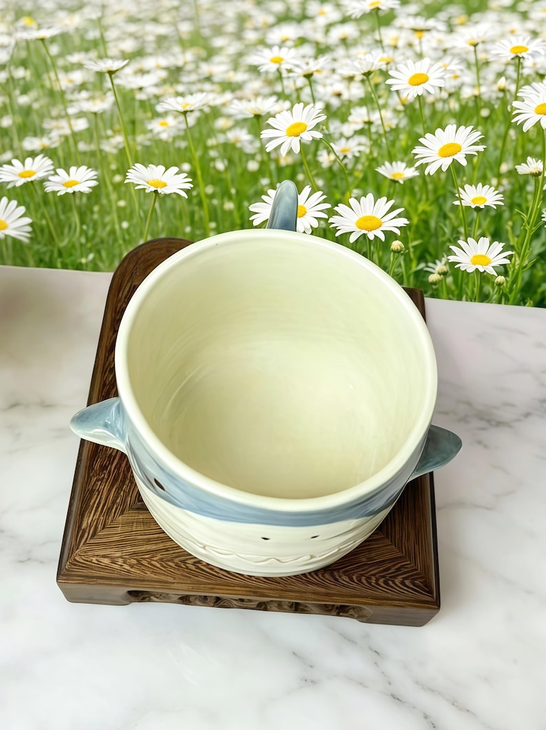 Puede incluir: Un cuenco de cer&aacute;mica con tem&aacute;tica de tibur&oacute;n con un dise&ntilde;o azul claro y blanco, con asa y aletas. El cuenco se encuentra sobre un soporte de madera oscura, con un fondo de margaritas.