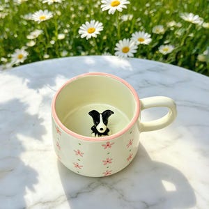 以下が含まれることがあります： クリーム色のセラミックマグカップ。ピンクの花柄とピンクの縁取りが施されています。内側には、小さな白黒の犬の置物があります。マグカップには取っ手があり、飲み物用である可能性があります。