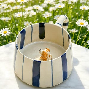 Puede incluir: Una taza de cer&aacute;mica con base color crema y rayas verticales azul marino. La taza cuenta con un asa curva y una peque&ntilde;a figura tridimensional de un perro marr&oacute;n y blanco adherida al fondo del interior del recipiente.