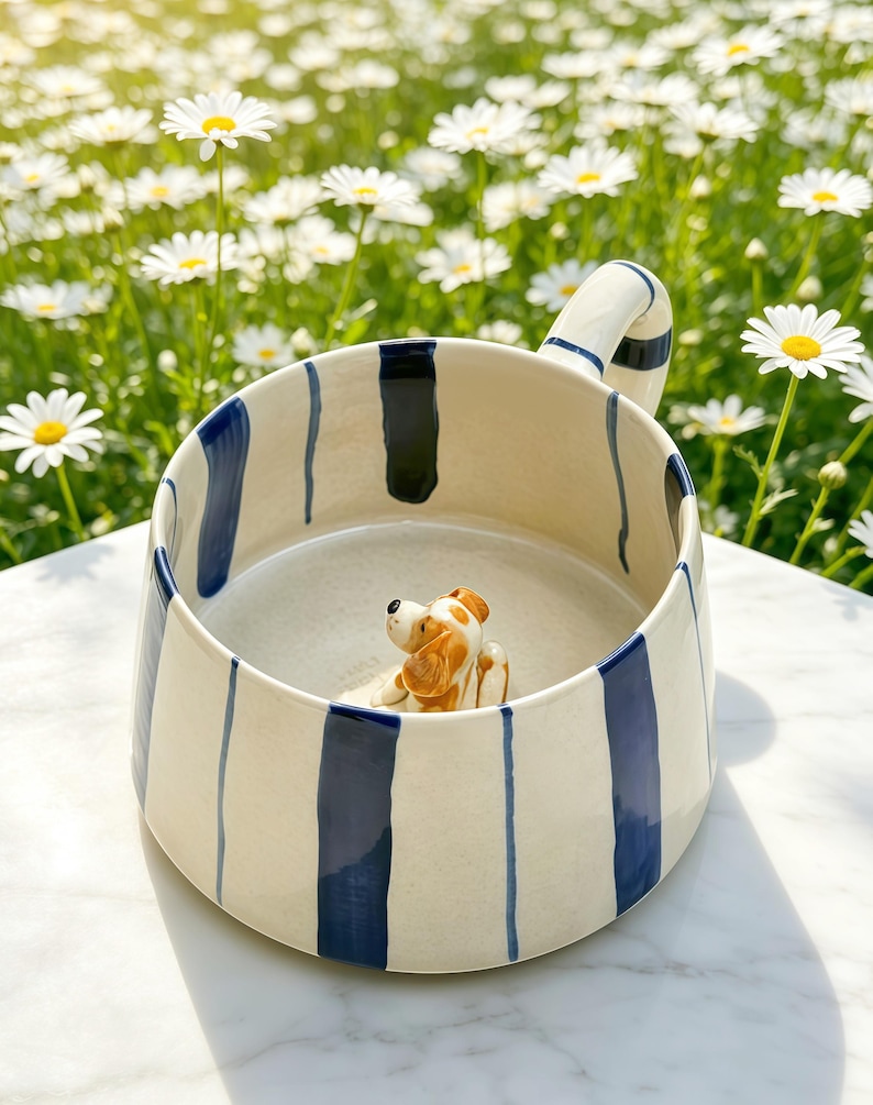 Puede incluir: Una taza de cer&aacute;mica con base color crema y rayas verticales azul marino. La taza cuenta con un asa curva y una peque&ntilde;a figura tridimensional de un perro marr&oacute;n y blanco adherida al fondo del interior del recipiente.