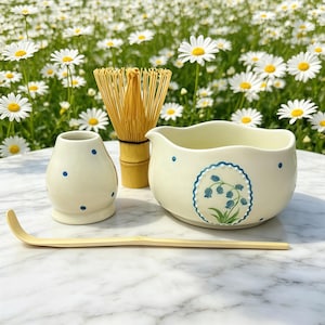 Puede incluir: Un juego de té matcha con un cuenco de cerámica color crema con un diseño floral azul, un jarrón pequeño a juego con lunares azules, un batidor de bambú y una cuchara de bambú. El juego es para preparar té matcha.