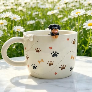 Könnte beinhalten: Ein cremefarbener Keramikbecher mit einer kleinen, dreidimensionalen schwarz-braunen Welpenfigur am Rand. Die Außenseite ist mit einem Muster aus schwarzen und braunen Pfotenabdrücken, orangefarbenen Knochen und kleinen roten Herzen sowie einem weißen Knochen verziert.