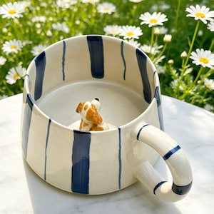 Puede incluir: Una taza de cer&aacute;mica con base color crema y rayas verticales azul marino. Dentro de la taza, una peque&ntilde;a figura de perro de cer&aacute;mica en tres dimensiones con manchas marrones y blancas est&aacute; fijada en el fondo, mirando hacia el borde superior.