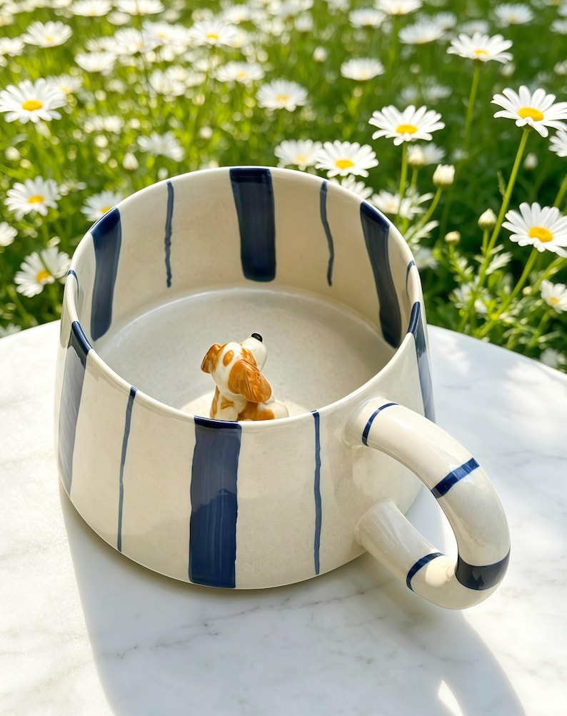 Puede incluir: Una taza de cer&aacute;mica con base color crema y rayas verticales azul marino. Dentro de la taza, una peque&ntilde;a figura de perro de cer&aacute;mica en tres dimensiones con manchas marrones y blancas est&aacute; fijada en el fondo, mirando hacia el borde superior.