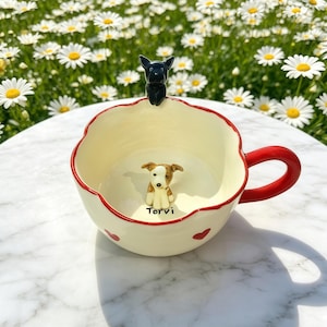 Könnte beinhalten: Eine cremefarbene Keramiktasse mit rotem Henkel und Rand, verziert mit roten Herzmotiven. Die Tasse hat eine kleine braun-weiße Hundefigur im Inneren und eine schwarze Hundefigur am Rand. Das Wort "Torvi" ist auf die Tasse gedruckt.