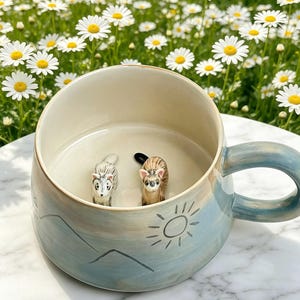 Puede incluir: Una taza de cerámica con exterior azul claro, con un diseño de montaña y sol pintado a mano. En el interior, se ven dos pequeñas figuras de hurones. La taza tiene un asa grande y está sobre un fondo de margaritas.