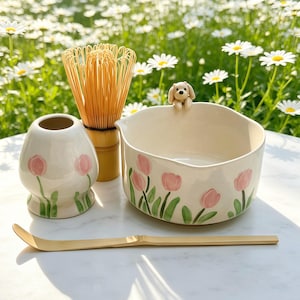 Op de afbeelding: Een keramische matchakom met een klein gebeeldhouwd hondje op de rand, een bijpassende gardehouder, een bamboe garde en een bamboe lepel. De kom en houder zijn voorzien van handgeschilderde roze tulpen met groene stelen op een crèmekleurige basis.