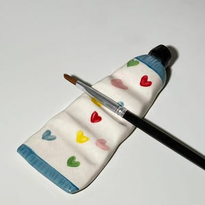 Può includere: Un portapennelli in ceramica a forma di tubetto di vernice con un pennello con manico nero. Il supporto è bianco con bordi blu e accenti a forma di cuore colorati. Il pennello poggia diagonalmente sul supporto.