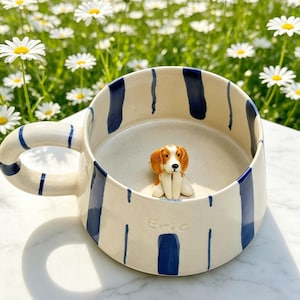 Peut inclure: Une tasse en céramique couleur crème ornée de rayures verticales bleu marine peintes et d'une figurine de chien marron et blanc sculptée et peinte à la main fixée au fond. Le nom Eric est gravé sur le côté extérieur de la tasse.