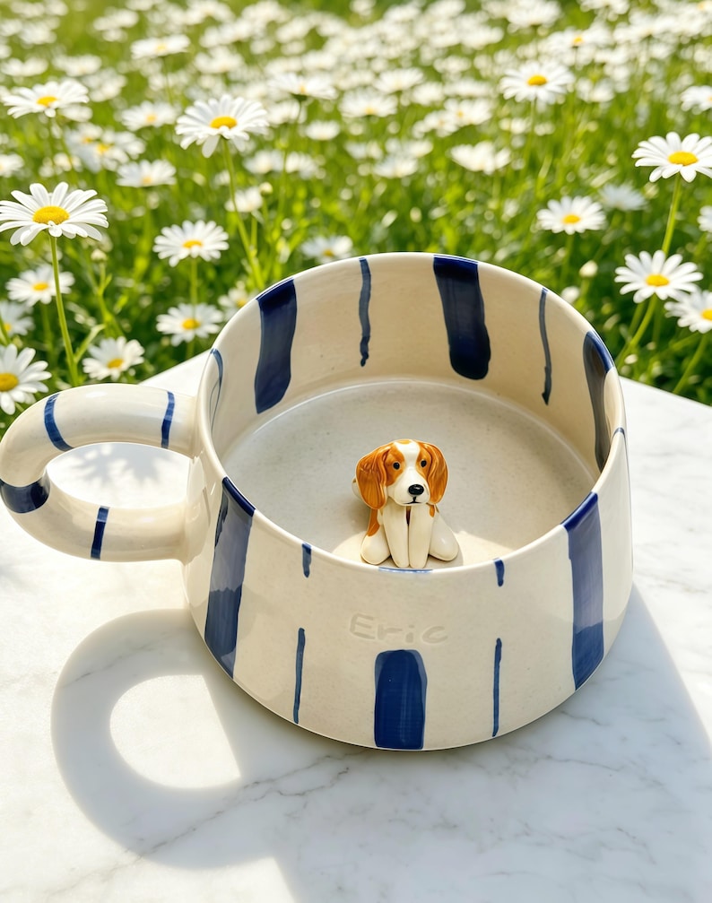 Puede incluir: Una taza de cer&aacute;mica color crema con rayas verticales azul oscuro y una figura esculpida de un perro marr&oacute;n y blanco en el fondo. El exterior de la taza tiene el nombre Eric grabado en relieve cerca de la base.