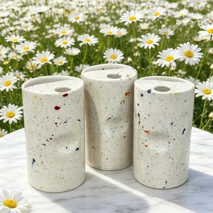 Puede incluir: Tres jarrones cilíndricos de cerámica blanca moteada, con un agujero en la parte superior. Los jarrones tienen salpicaduras de pintura de colores y están colocados sobre una mesa de mármol. El fondo presenta un campo de margaritas.