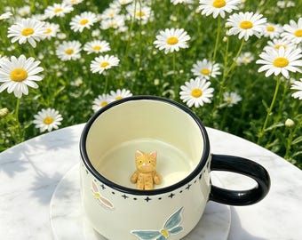Puede incluir: Una taza de cerámica color crema con asa y borde negros, decorada con calcomanías de mariposas y una pequeña figura de gato en el interior. La taza está sobre un posavasos de mármol, con un fondo de margaritas.