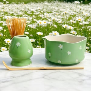 Op de afbeelding: Een matcha theeset met een groene keramische kom en klopperhouder, beide versierd met witte bloemmotieven. Een bamboe klopper en lepel zijn ook inbegrepen. De set is ontworpen voor het bereiden van matcha thee.