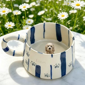 Puede incluir: Una taza de cer&aacute;mica color crema con rayas verticales azul oscuro, huellas de patas grises y el nombre Liam escrito en gris. Una peque&ntilde;a figura de perro esculpida en tres dimensiones est&aacute; fijada en la base interior de la taza.