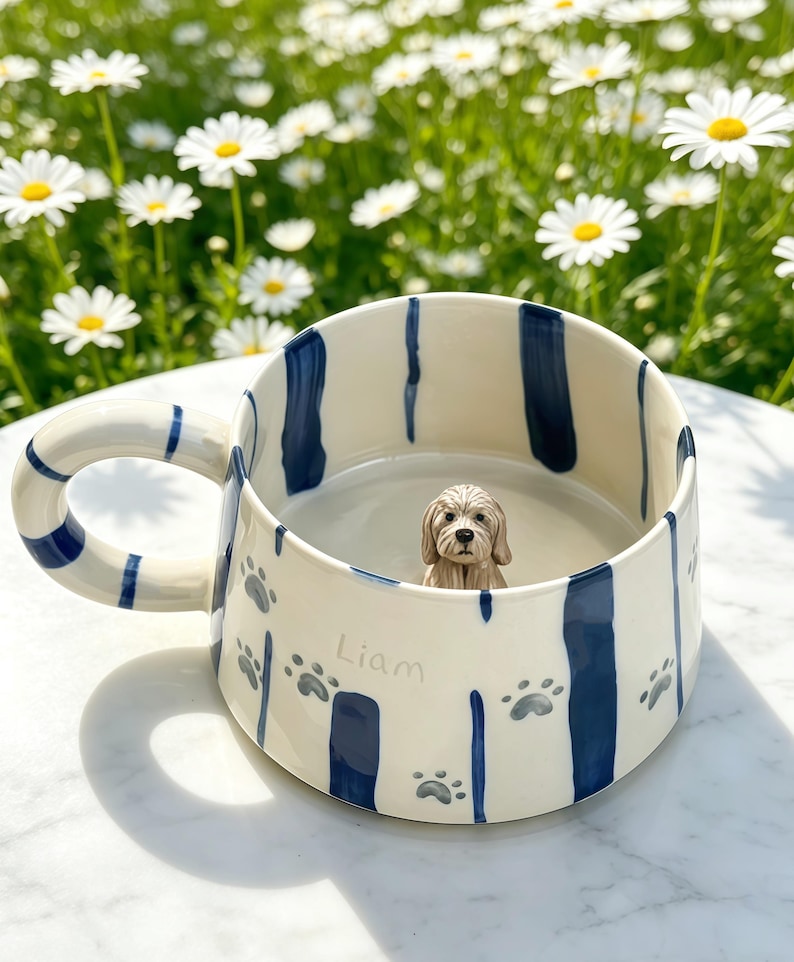 Puede incluir: Una taza de cer&aacute;mica color crema con rayas verticales azul oscuro, huellas de patas grises y el nombre Liam escrito en gris. Una peque&ntilde;a figura de perro esculpida en tres dimensiones est&aacute; fijada en la base interior de la taza.