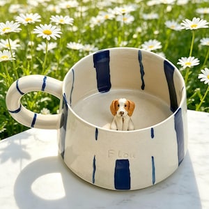 Puede incluir: Una taza de cer&aacute;mica color crema con rayas verticales pintadas en azul oscuro y un asa grande. Dentro de la taza hay una peque&ntilde;a figura tridimensional de un perro marr&oacute;n y blanco. La palabra "Flou" est&aacute; grabada en la superficie exterior.