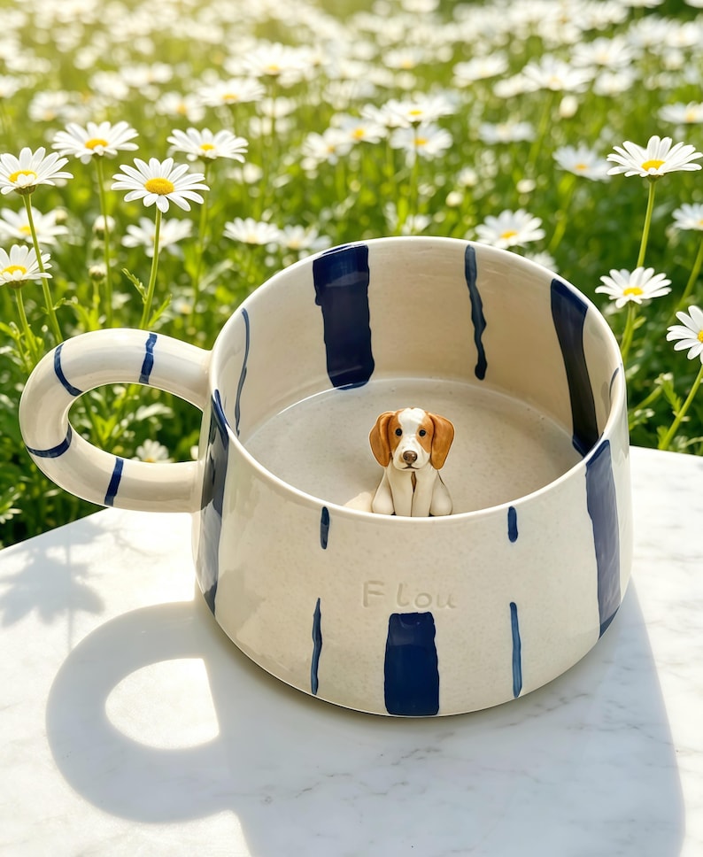 Puede incluir: Una taza de cer&aacute;mica color crema con rayas verticales pintadas en azul oscuro y un asa grande. Dentro de la taza hay una peque&ntilde;a figura tridimensional de un perro marr&oacute;n y blanco. La palabra "Flou" est&aacute; grabada en la superficie exterior.
