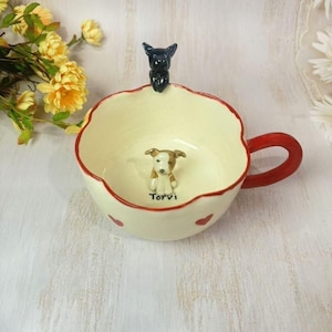 May include: A ceramic teacup with a red handle and rim, featuring a small dog figurine inside and a black dog figurine on the rim. The cup has a cream-colored interior and is decorated with red hearts and the name "Torvi".