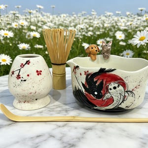 Puede incluir: Un juego de té matcha con un cuenco de cerámica blanca con un diseño de peces koi, un batidor de bambú, un chawan y una cuchara de bambú. El cuenco tiene dos pequeñas figuras de animales en el borde. El juego está sobre una superficie de mármol.