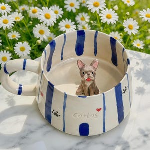 Puede incluir: Una taza de cer&aacute;mica con base color crema y rayas azules verticales. El exterior muestra el nombre Carlos, peque&ntilde;as huellas negras y corazones rojos. Dentro de la taza, una peque&ntilde;a figura tridimensional de un bulldog franc&eacute;s marr&oacute;n.