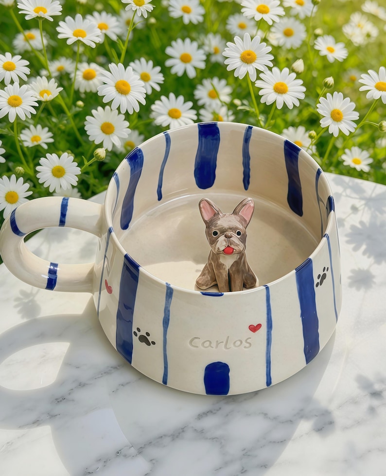 Puede incluir: Una taza de cer&aacute;mica con base color crema y rayas azules verticales. El exterior muestra el nombre Carlos, peque&ntilde;as huellas negras y corazones rojos. Dentro de la taza, una peque&ntilde;a figura tridimensional de un bulldog franc&eacute;s marr&oacute;n.