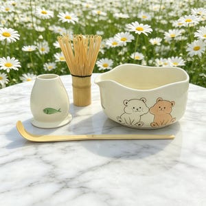 May include: A matcha tea set includes a ceramic bowl with bear illustrations, a bamboo whisk, a bamboo scoop, and a ceramic water container with a fish design. The set is cream-colored and sits on a marble surface.