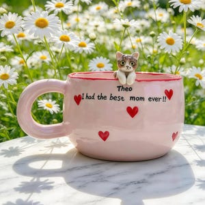 Può includere: Una tazza in ceramica rosa chiaro con una piccola figura di gatto scolpita appollaiata sul bordo. La tazza è decorata con diversi piccoli cuori rossi e il testo nero "Theo I had the best mom ever!!" stampato sulla parte anteriore.