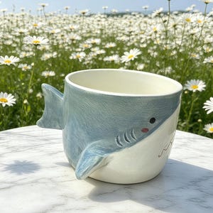 Puede incluir: Una taza de cer&aacute;mica con forma de tibur&oacute;n, con exterior azul claro e interior blanco. La taza tiene un asa de aleta y una cara pintada con ojos negros y boca roja. La taza est&aacute; sobre una superficie blanca.