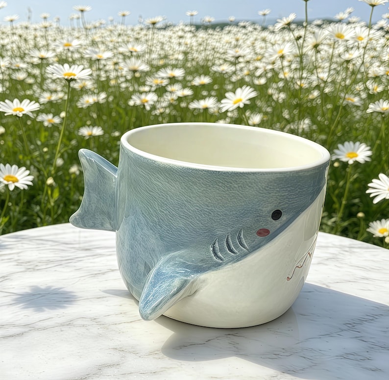 Puede incluir: Una taza de cer&aacute;mica con forma de tibur&oacute;n, con exterior azul claro e interior blanco. La taza tiene un asa de aleta y una cara pintada con ojos negros y boca roja. La taza est&aacute; sobre una superficie blanca.