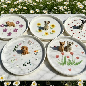 Puede incluir: Una colección de cinco platos de cerámica blanca, cada uno adornado con una figura de perro en miniatura y diseños florales. Los platos presentan varios colores, incluyendo rosa, amarillo y rojo. Los platos están dispuestos sobre una superficie de mármol.