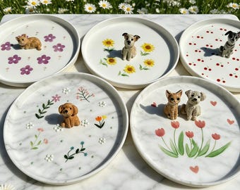 Puede incluir: Una colección de cinco platos de cerámica blanca, cada uno adornado con una figura de perro en miniatura y diseños florales. Los platos presentan varios colores, incluyendo rosa, amarillo y rojo. Los platos están dispuestos sobre una superficie de mármol.