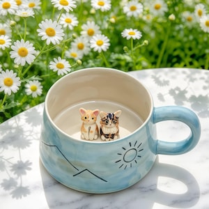 Puede incluir: Una taza de cerámica azul claro con acabado brillante, que presenta un dibujo lineal negro de un sol y una cordillera en el exterior. Dentro de la taza, dos pequeñas figuras de gato detalladas están fijadas en la base, una naranja y blanca y otra atigrada marrón y negra.