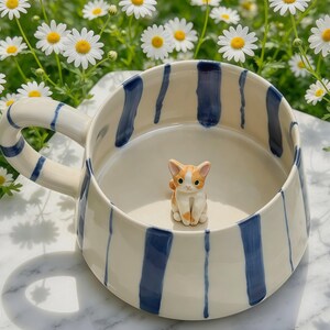 Puede incluir: Una taza de cer&aacute;mica con base color crema y rayas verticales azul marino. La taza cuenta con un asa ancha y una peque&ntilde;a figura tridimensional de un gato naranja y blanco fijada en el centro de la base interior del recipiente.