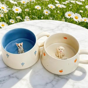 Può includere: Due tazze in ceramica con manici. La tazza a sinistra ha un interno blu con una piccola statuetta di gatto e fiori blu sull'esterno color crema. La tazza a destra ha un interno color crema con una piccola statuetta di cane e pesche.