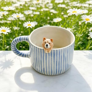 Może przedstawiać: Ceramiczny kubek w pionowe jasnoniebieskie paski z małą, trójwymiarową figurką psa w kolorze brązowo-białym przymocowaną do wewnętrznej krawędzi. Kubek posiada pasujący wzorzysty uchwyt oraz gładką, kremową powierzchnię wewnętrzną.
