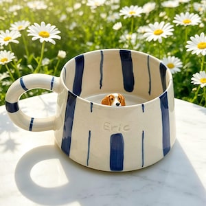 Puede incluir: Una taza de cer&aacute;mica color crema con rayas verticales azul marino y un asa curva. El nombre Eric est&aacute; grabado en la parte frontal. Una peque&ntilde;a figura tridimensional de un perro marr&oacute;n y blanco est&aacute; fijada en la base interior de la taza.
